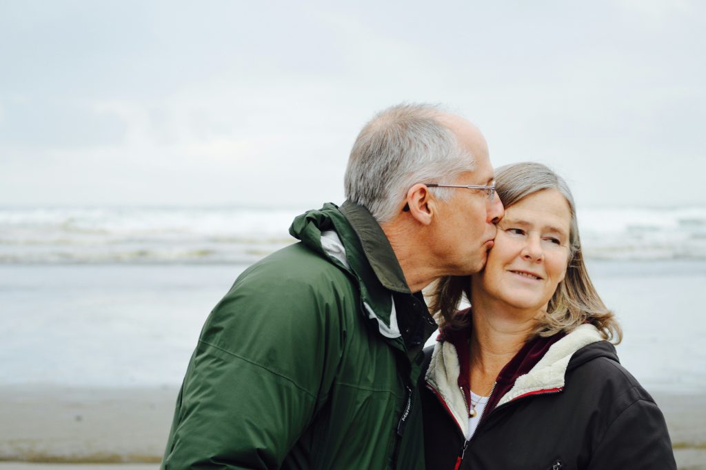 an elderly couple on the beach. the man is kissing the woman on the cheek