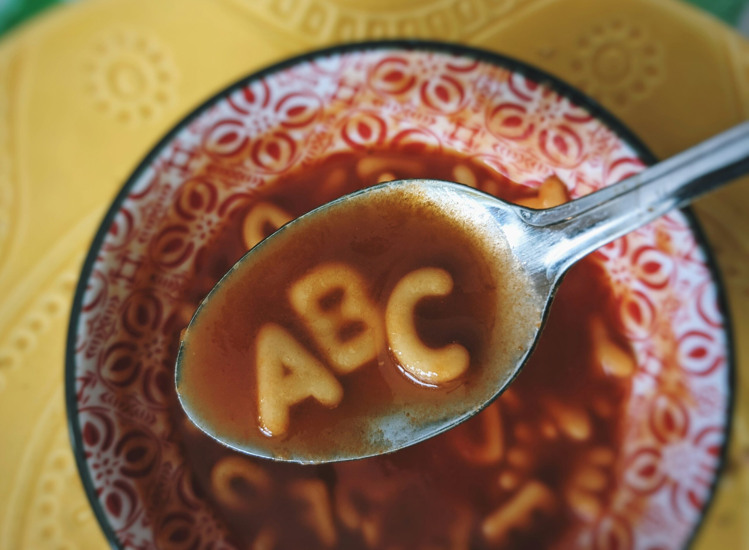 a spoon held above a bowl of red soup holds noodles in the shape of A, B, C.